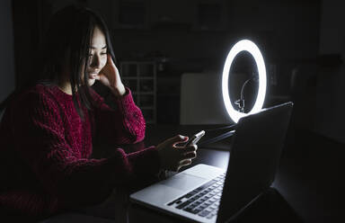 Junge Influencerin, die ihr Smartphone benutzt, während sie mit ihrem Laptop vor dem Ringlicht in der Dunkelkammer sitzt - LJF02098