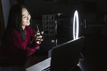 Lächelnde Vloggerin, die ihr neues Smartphone vor einem Ringlicht hält, während sie sendet - LJF02107