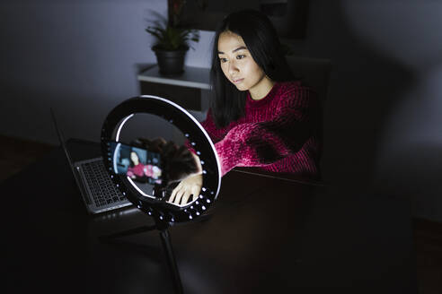 Young Chinese female vlogger sitting with laptop and illuminated ring light - LJF02108