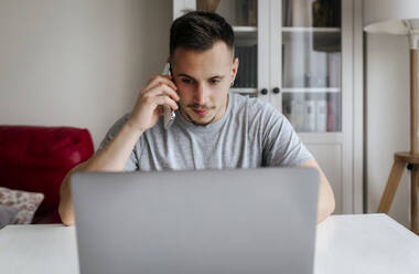Junger männlicher Unternehmer, der mit einem Smartphone spricht, während er im Büro zu Hause einen Laptop benutzt - MGOF04663