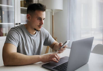 Handsome male entrepreneur with smart phone using laptop on table at home office - MGOF04664