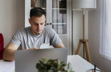 Young male entrepreneur using smart phone while sitting with laptop at home office - MGOF04666