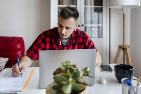 Gut aussehender männlicher Unternehmer mit In-Ear-Kopfhörern beim Schreiben am Laptop im Home Office - MGOF04673