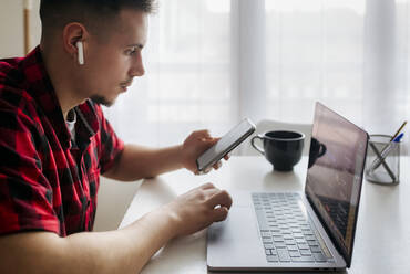 Männlicher Unternehmer mit In-Ear-Kopfhörern, der sein Smartphone in der Hand hält, während er im Heimbüro am Laptop arbeitet - MGOF04675
