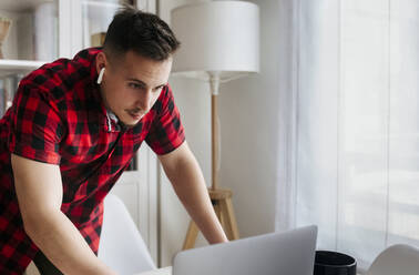 Male entrepreneur with in-ear headphones leaning on table while using laptop at home office - MGOF04677