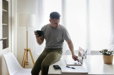 Geschäftsmann hält eine Kaffeetasse und benutzt einen Laptop, während er im Büro zu Hause auf dem Schreibtisch sitzt - MGOF04680