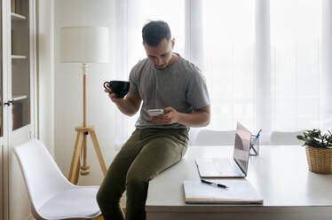 Junger männlicher Unternehmer, der einen Kaffee trinkt und dabei sein Smartphone benutzt, während er im Büro zu Hause am Schreibtisch sitzt - MGOF04681