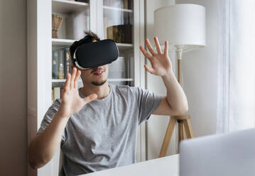 Young man gesturing while wearing Virtual reality headset at home office - MGOF04687