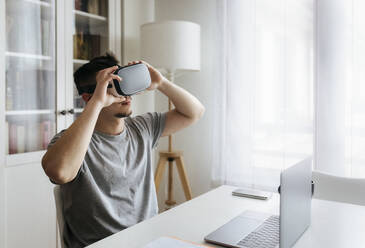 Male entrepreneur holding Virtual reality headset while sitting with laptop on desk at home office - MGOF04688