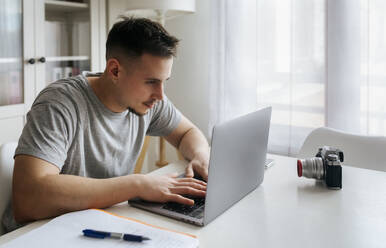 Junger männlicher Berufstätiger, der einen Laptop benutzt, während er ein Video mit einer Digitalkamera im Heimbüro aufnimmt - MGOF04693