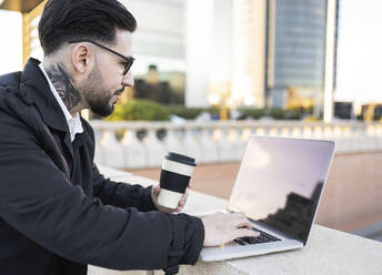 Geschäftsmann hält Kaffeetasse, während er einen Laptop an einer Stützmauer benutzt - JCCMF01374