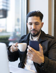 Männlicher Unternehmer hält Kaffeetasse während Videoanruf in Café - JCCMF01377