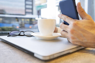 Die Hand eines Geschäftsmannes, der ein Smartphone benutzt, während er einen Kaffee mit einem Laptop am Kaffeetisch trinkt - JCCMF01378