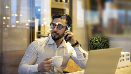 Businessman talking on smart phone while holding coffee cup in cafe - JCCMF01379