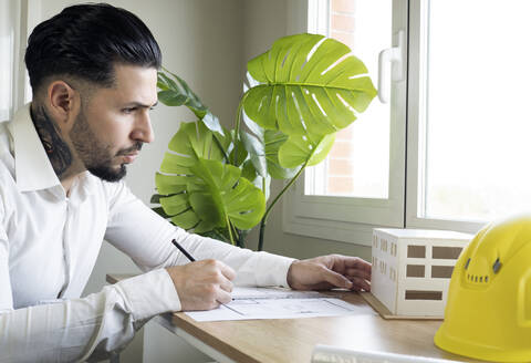 Männlicher Architekt, der ein architektonisches Modell betrachtet, während er einen Bauplan im Büro zeichnet - JCCMF01384