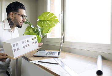 Männlicher Architekt, der ein Architekturmodell während eines Videogesprächs über einen Laptop am Schreibtisch hält - JCCMF01390
