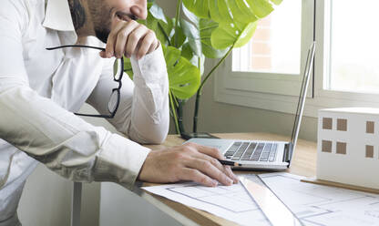 Male design professional holding eyeglasses while using laptop at desk in office - JCCMF01392