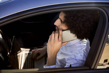Männlicher Unternehmer, der mit seinem Smartphone telefoniert, während er einen Laptop im Auto benutzt - VEGF04050