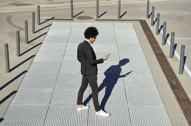 Männlicher Berufstätiger, der ein Smartphone benutzt, während er an einem sonnigen Tag auf dem Gehweg steht - VEGF04054