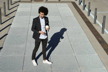 Male entrepreneur holding coffee cup and mobile phone while standing on footpath - VEGF04055