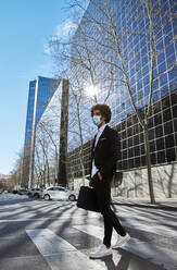 Businessman with laptop bag walking on street against building during sunny day during COVID-19 - VEGF04056