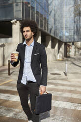 Geschäftsmann mit Laptoptasche und Kaffeetasse beim Überqueren der Straße an einem sonnigen Tag - VEGF04070
