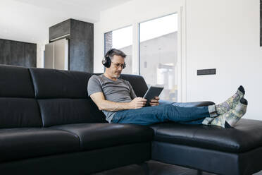 Älterer Mann hört Musik über Kopfhörer, während er ein digitales Tablet im Wohnzimmer benutzt - JRFF05102