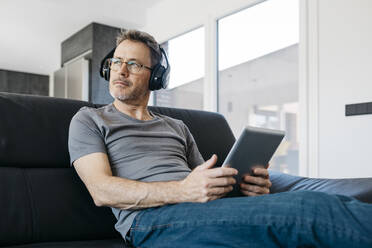 Mature man with digital tablet looking away while listening music through headphones on sofa in living room - JRFF05104