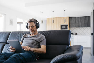 Älterer Mann mit Kopfhörern, der ein digitales Tablet benutzt, während er zu Hause auf dem Sofa sitzt - JRFF05105