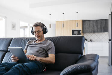 Mature man with headphones using digital tablet while sitting on sofa at home - JRFF05105