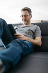 Lächelnder reifer Mann, der ein digitales Tablet benutzt, während er auf dem Sofa sitzt - JRFF05107