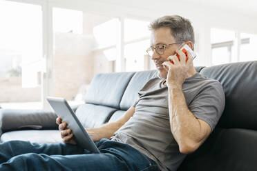 Älterer Mann, der auf dem Sofa im Wohnzimmer mit einem Mobiltelefon spricht und ein digitales Tablet benutzt - JRFF05108