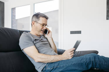 Älterer Mann, der auf dem Sofa im Wohnzimmer mit einem Smartphone spricht und ein digitales Tablet benutzt - JRFF05109