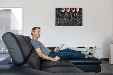 Mature man with digital tablet looking away while sitting on sofa at home - JRFF05110