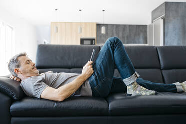 Mature man watching at digital tablet while lying down on sofa in living room - JRFF05111