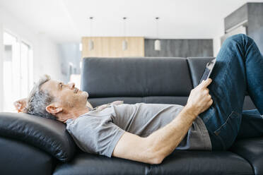 Smiling mature man with digital tablet day dreaming while lying down on sofa in living room - JRFF05112