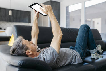 Mature man using digital tablet while lying on sofa at living room - JRFF05113