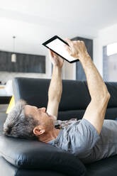 Älterer Mann benutzt ein digitales Tablet, während er auf dem Sofa im Wohnzimmer liegt - JRFF05114