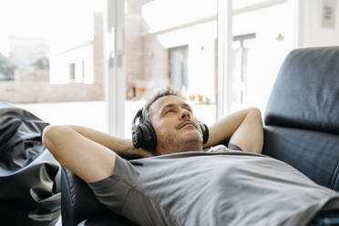 Älterer Mann hört Musik über Kopfhörer, während er auf dem Sofa im Wohnzimmer liegt - JRFF05115