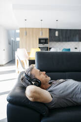 Mann mit Händen hinter dem Kopf hört Musik über Kopfhörer, während er sich auf dem Sofa zu Hause entspannt - JRFF05117