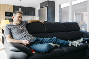 Mature man holding remote control while sitting on sofa at home - JRFF05121
