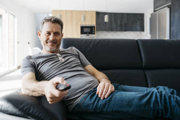 Smiling man holding TV remote while sitting on sofa at living room - JRFF05123