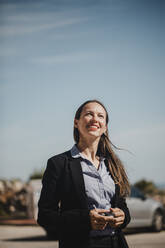 Smiling businesswoman looking up while standing against sky on sunny day - DMGF00533