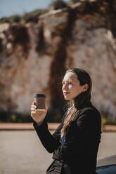 Nachdenkliche Geschäftsfrau schaut weg, während sie eine Einweg-Kaffeetasse an einem sonnigen Tag hält - DMGF00539