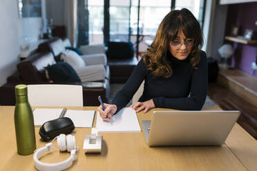 Mature woman writing in paper while sitting by laptop at home - EGAF02015