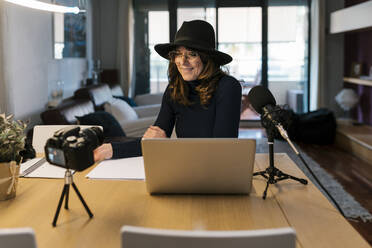 Mature influencer with laptop and microphone stand smiling while filming on camera at home - EGAF02027