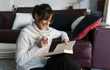 Frau mit Kaffeetasse lächelnd beim Lesen eines Buches zu Hause - EGAF02033