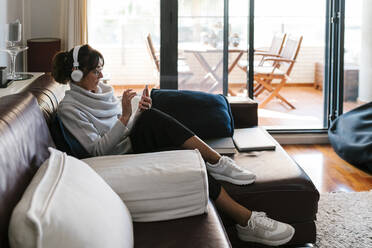 Reife Frau mit Kopfhörern, die ein Mobiltelefon benutzt, während sie zu Hause sitzt - EGAF02034