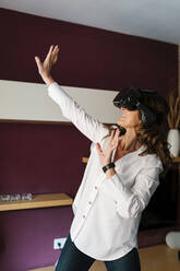 Mature woman with virtual reality headset stretching hands while playing at home - EGAF02041