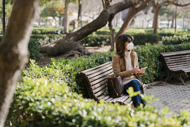 Mature woman with protective face mask holding mobile phone while sitting at park - EGAF02043